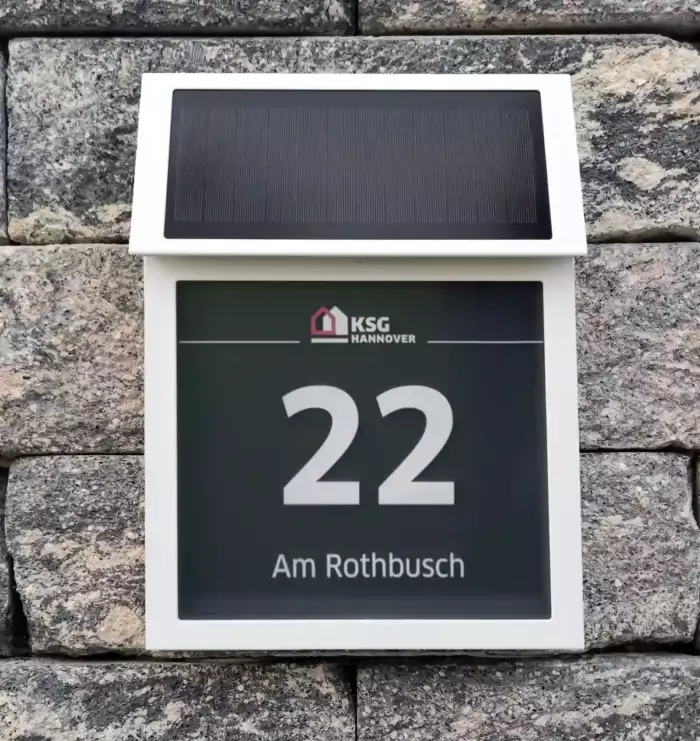 Solar-Hausnummer mit weißem Gehäuse. Das quadratische Feld hat die Aufschrift Hausnummer 22 und Straße Am Rothbusch und sowie das Logo KSG Hannover darüber. Die Hintergrundfarbe des Feldes ist grau.